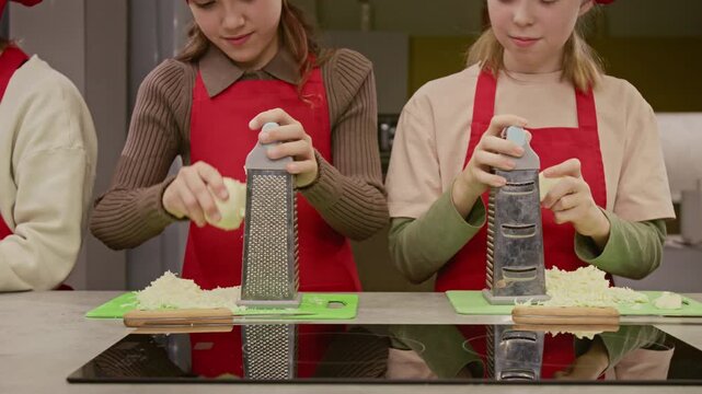 Caucasian teenage girls wearing red aprons grating onions on metal graters during cooking class. Footage suitable for promoting culinary schools or children cooking workshops