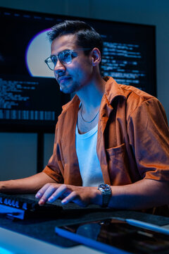 A serious young cyber threat analyst points a finger while looking at the screen in a dimly lit network monitoring room, under the blue glow of security dashboards.