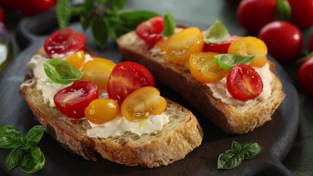 Close-up horizontal video of a hand putting basil leaves on toasted Italian bread, or bruschetta, with creamy stracciatella cheese and red and yellow cherry tomatoes. Mediterranean appetizer, homemade