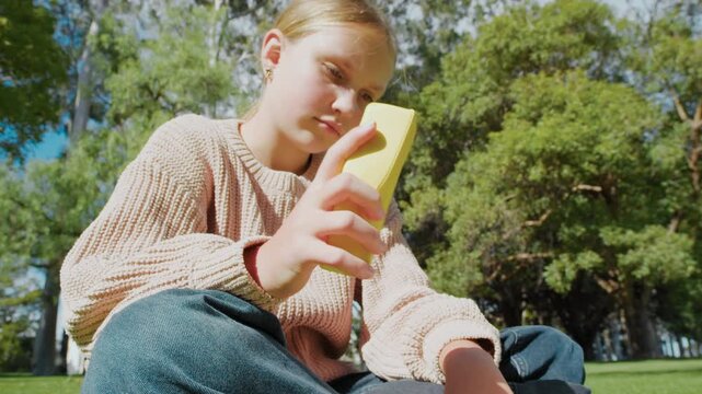Close-up of a young girl sits on the grass in a park. She reads a book and plays with a squishy toy and focuses on squeezing it.
