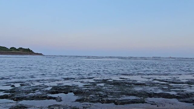 sea and sky Muted blues frame calm coastal tide pools. Shallow water reveals textured, wet rocks. Faint horizon shows distant green shoreline. Quiet, serene scene at low tide. Perfect for tranquil