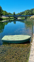 Fototapeta premium A small green rowboat is tied near the shore of the calm Dobra river in Jarce Polje with a concrete road bridge in the background under a blue sky