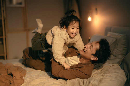 Asian middle aged man lying on bed lifting Asian child in air, both smiling and playing together in cozy bedroom during evening, family bonding and spending quality time at home