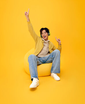 Asian man sitting on a beanbag with headphones and pointing upward expressing music enjoyment youthful energy digital entertainment creative freedom and a cheerful modern lifestyle.