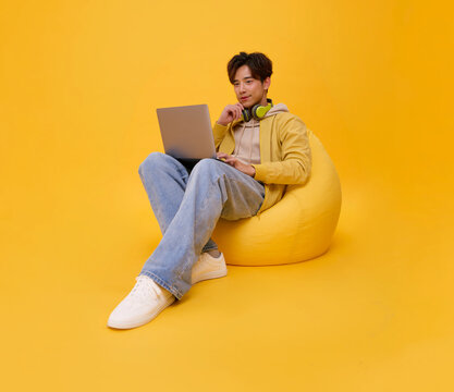 Asian man using a laptop while relaxing on a beanbag with headphones representing remote work online learning digital creativity flexible productivity and a modern connected lifestyle.