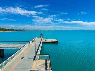 Obraz premium Clear blue water meets a bright sky at a pier with concrete pathways and metal railings near a sandy beach in a coastal area during daytime