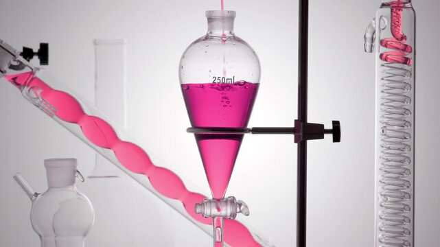 Lab setup with clear pink transparent liquid poured in a thin stream into a separatory funnel, making bubbles underwater and flowing down a lab condenser coil on a smooth grey ramp bg
