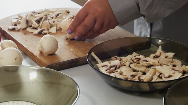 A woman's hands slice champignons with a knife on a cutting board on a table with vegetables. Medium shot.