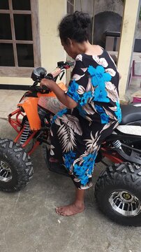 Barefoot Asian woman in a floral dress checking the controls of an orange ATV parked in a residential courtyard