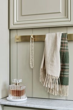 Closeup of a brushed brass hook rail displaying a cream scarf and a green plaid scarf, with a decorative glass bottle on a marble surface in a stylish interior setting