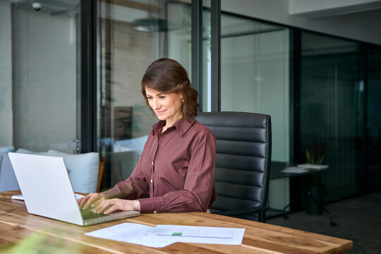 Older mature businesswoman working on laptop computer in office. Busy woman financial director checks corporate email online. Female business executive manages company at workplace. Copy space