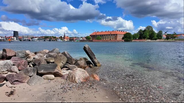 View across Flensburg Fjord toward the historic Sonderborg with clear water, a rocky shoreline, sandy beach and Mill in the background.