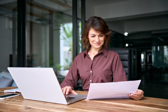 Latin businesswoman checking paper report at desk in office with copy space. Mature woman financial analyst holds document, works on laptop. Female expert reviews business contract at workplace.