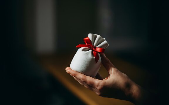 A person holds a small gift wrapped in white fabric tied with a red ribbon on a wooden surface in soft lighting