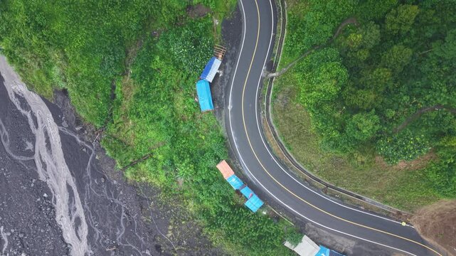Aerial drone photograph of a winding mountain road curving beside a rocky riverbed, with temporary tarpaulin shelters tucked into lush green vegetation, highlighting remote infrastructure