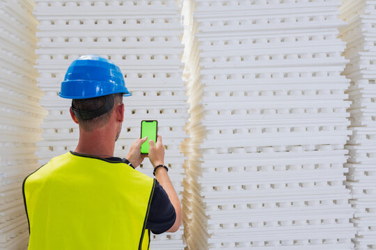 Back view of a worker with a blue hard hat using a green screen smartphone between stacks of polystyrene insulation