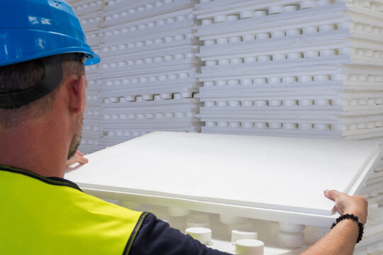 Worker in a blue hard hat and yellow safety vest carrying a white polystyrene insulation panel