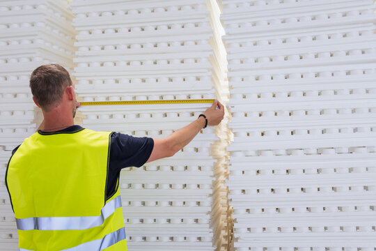 Back view of a construction worker in a high-visibility vest measuring a stack of white polystyrene insulation sheets