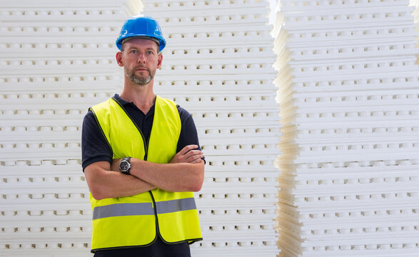 Portrait of a confident construction worker in a blue hard hat standing with crossed arms by polystyrene insulation sheets
