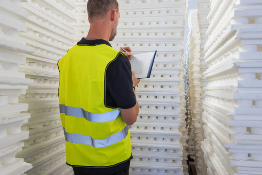Back view of a warehouse inspector in a high-visibility vest writing on a clipboard among stacks of polystyrene sheets