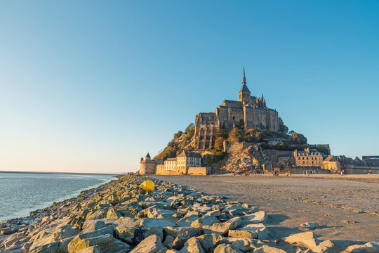 Mont Saint Michel abbey above rocky shoreline at sunset with medieval French architecture and scenic coastal landscape in Normandy, France. Tidal island with monastery in Normandie. Coastal landscape
