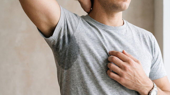A close-up of a person experiencing excessive underarm sweating, a large visible sweat stain on a light gray T-shirt. One arm is raised to clearly reveal the damp armpit area.