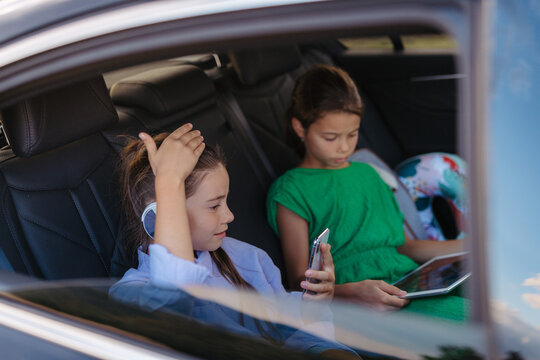 Little sisters sitting in car, playing on tablet and smartphone.