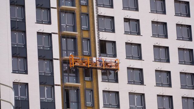 Construction workers on suspended platform installing facade of modern apartment building, exterior renovation and urban residential development with windows and insulation system.