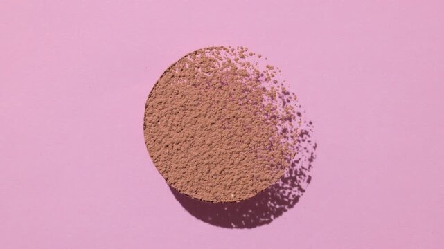 From-above shot of brown powder sifted in the form of a circle creates a shifting shadow pattern on a light pink backdrop