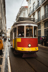 Fototapeta premium Traditional red Lisbon tram traveling through the historic city center on a rainy day, surrounded by classic Portuguese architecture and lively urban activity.
