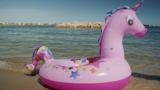 Pink unicorn inflatable float resting on the sandy shore of Plage de la Gravette in Antibes, France, with calm Mediterranean water and rocky breakwater in the background