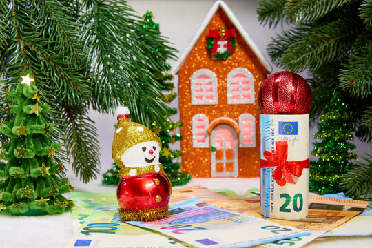 A decorated euro banknote and a snowman stand on top of euro banknotes against a backdrop of Christmas trees and an orange house