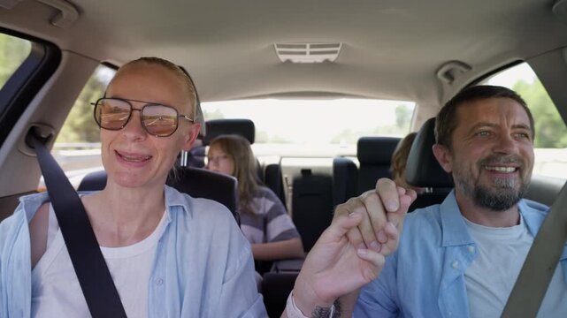 Happy family enjoying road trip inside modern car. Loving parents holding hands and smiling at each other while teenage son and daughter riding in back seats, family bonding and travel lifestyle.
