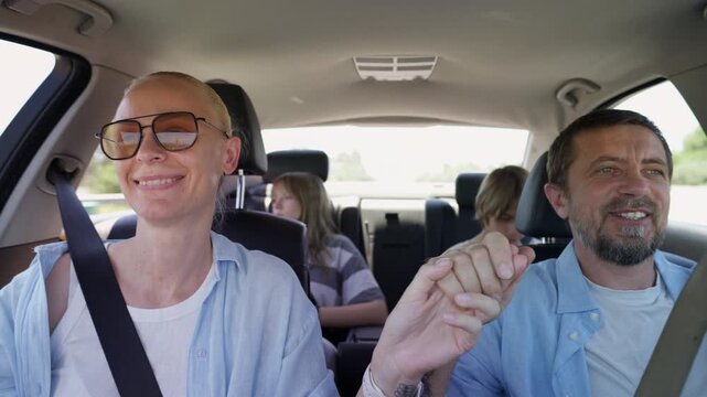Happy family traveling by car on summer road trip. Parents sharing loving glance and holding hands while teenage son and daughter relaxing in back seats, celebrating family connection and togetherness