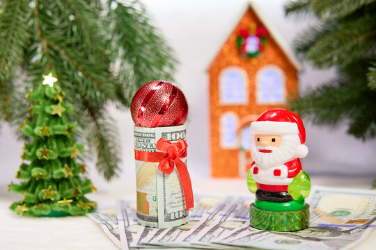 Santa Claus with a decorated dollar bill and a ball against the background of Christmas trees with a house