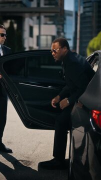 Caucasian bodyguard opening luxury car door for African American businessman stepping out on city street, vertical. Video is useful for executive security and VIP transport concepts