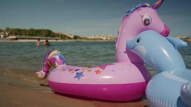 Colorful unicorn and dolphin inflatable floats on the shallow water at Plage de la Gravette with families swimming and the rocky breakwater in the background