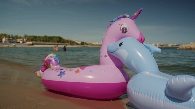 Colorful unicorn and dolphin inflatable floats on the shallow water at Plage de la Gravette with families swimming and the rocky breakwater in the background