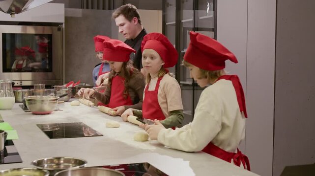 Caucasian children in chef hats rolling dough during culinary class while young instructor assists and child records video on smartphone. Ideal for promoting kids cooking workshops