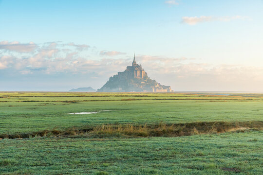 Mont Saint-Michel abbey rises above wide coastal marshes under morning light in Normandy, France. French medieval monastery at sunrise. Rural landscape, nature tourism, heritage travel