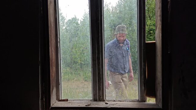 Bearded traveler walking and peering inside through a dirty window of an abandoned house
