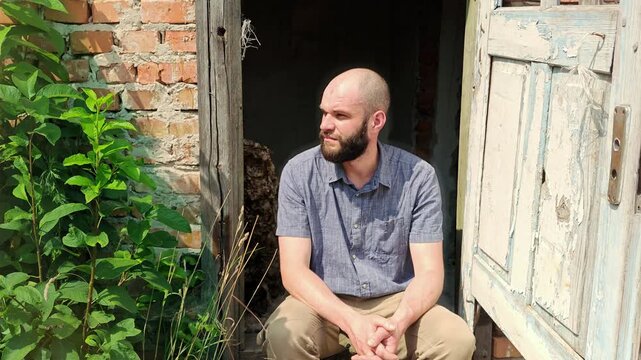Tired male traveler sitting on the doorstep of an abandoned brick building and resting