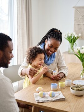 Happy African American family painting Easter eggs together at a wooden table. A smiling mother helps her young daughter decorate eggs with pastel colors in a bright, modern home setting