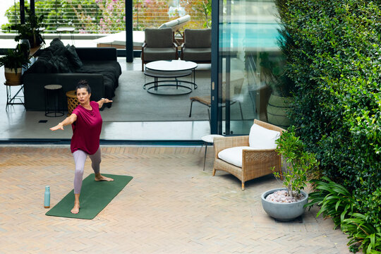 Mature woman practicing yoga on green mat on patio wearing leggings, water bottle nearby