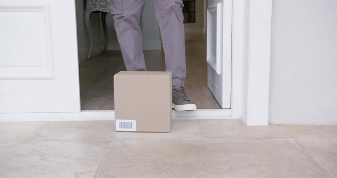 African American man spotting package at door, nudging and lifting it inside holding smartphone