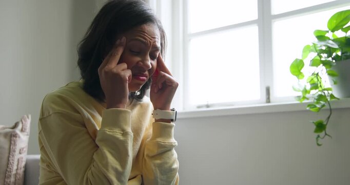 Adult African woman feeling headache, pressing temples, wincing and gazing by window, copy space