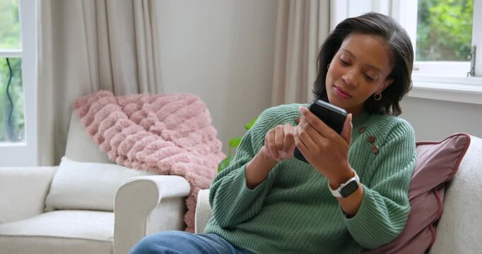 Woman in green sweater sitting on sofa near window holding phone, reading and replying to messages