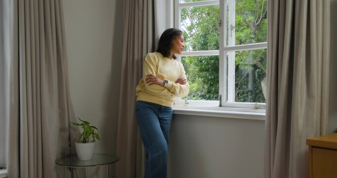 African American woman at home gazing at garden, uncrossing arms, placing hands on sash window sill