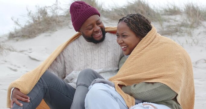 African American couple sitting on dune, man draping mustard blanket, seeking warmth and closeness