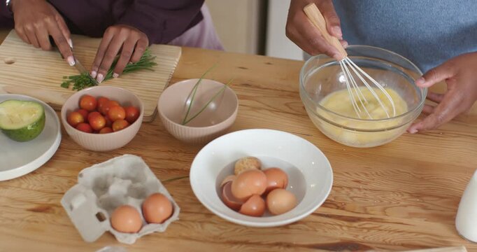 Couple preparing meal, whisking eggs in glass bowl while chopping herbs on wooden cutting board